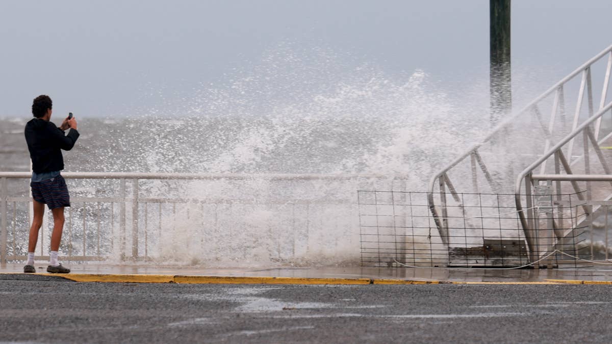 Man in shorts and jacket films 10-foot waves crashing near a pier on the Oregon Coast.