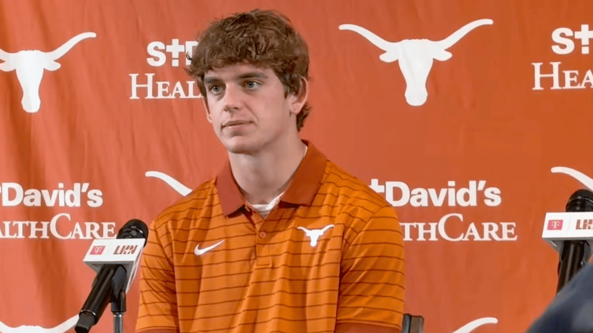 Arch Manning in an orange Texas polo shirt at a press conference with microphones and a St. David's HealthCare backdrop.