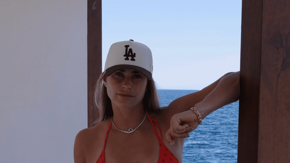 Aryna Sabalenka in red swimsuit and LA Dodgers hat poses by the sea.