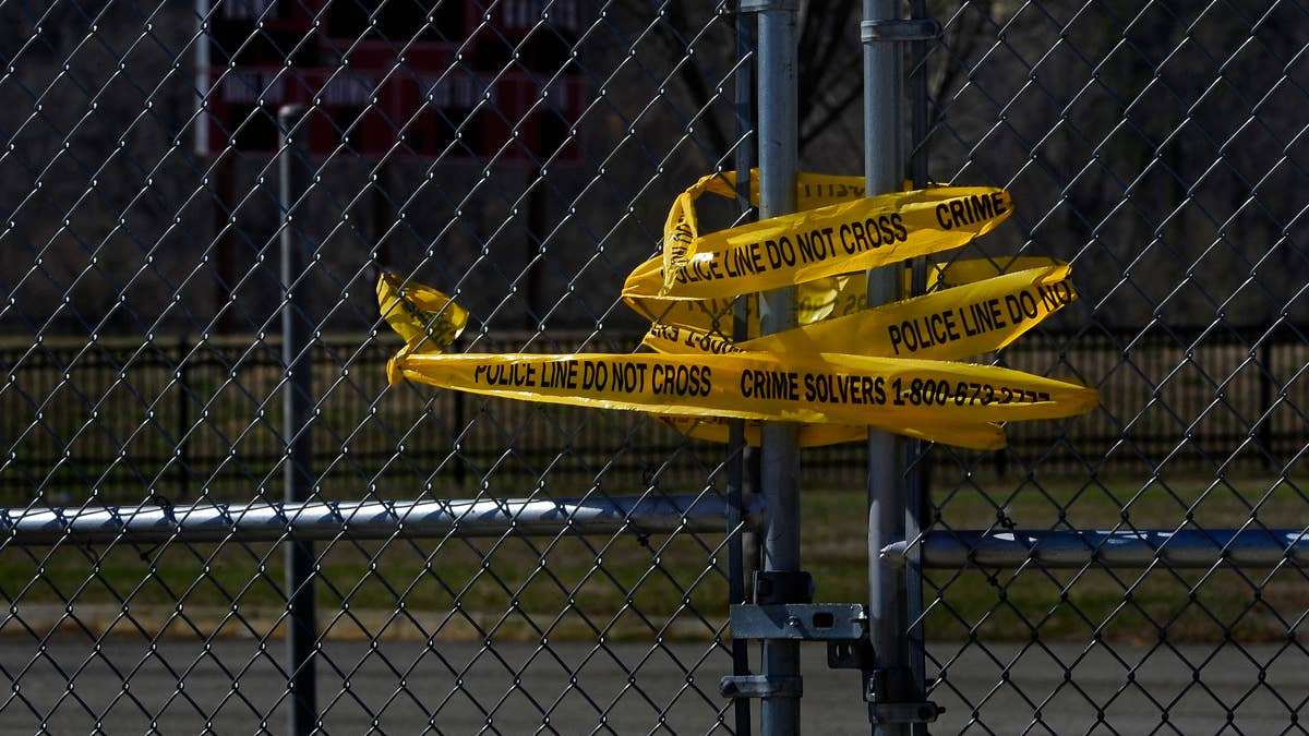 Yellow police tape reading "POLICE LINE DO NOT CROSS" and "CRIME SOLVERS 1-800-577-TIPS" is wrapped around a chain-link fence.