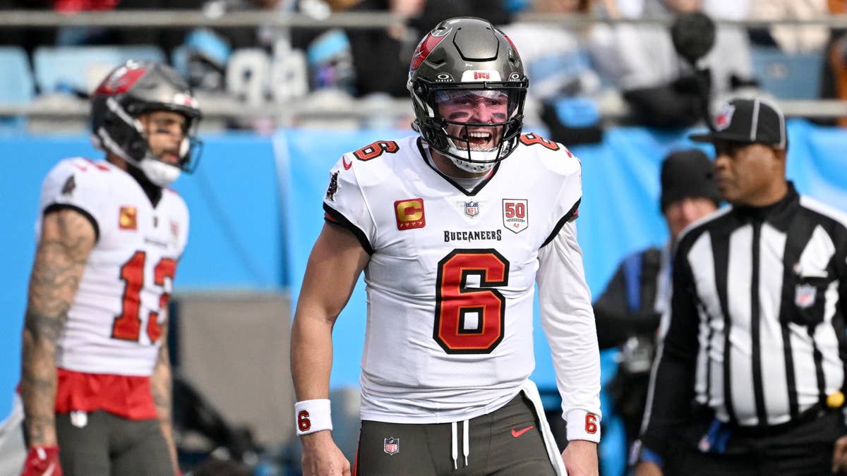 Baker Mayfield in a white Buccaneers jersey with number 6, on the field with a teammate and referee.