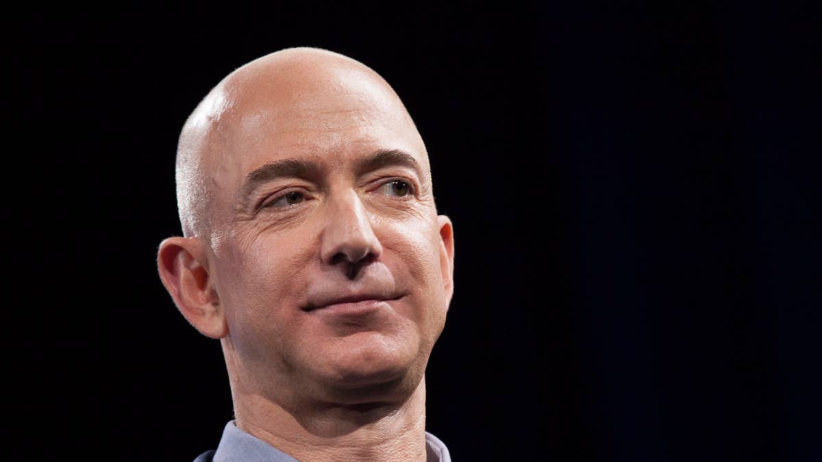 Jeff Bezos smiles, wearing a blue collared shirt against a dark background.
