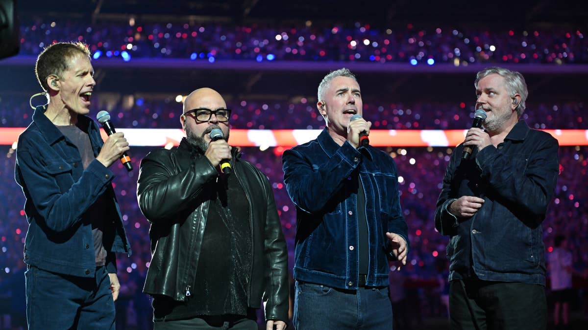 Four men singing into microphones on a stage with a blurred crowd in the background.
