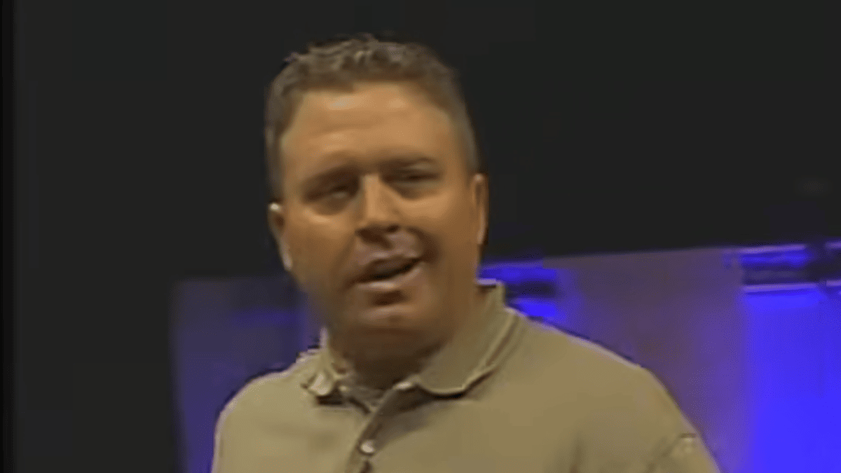 Randy Pedersen in a tan polo shirt speaking on stage with blue lighting in the background.