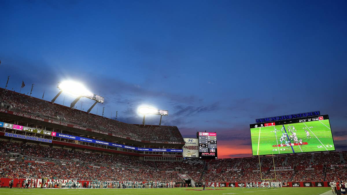 Raymond James Stadium with a large screen showing a Bucs-Browns game during a sunset and stadium lights on.
