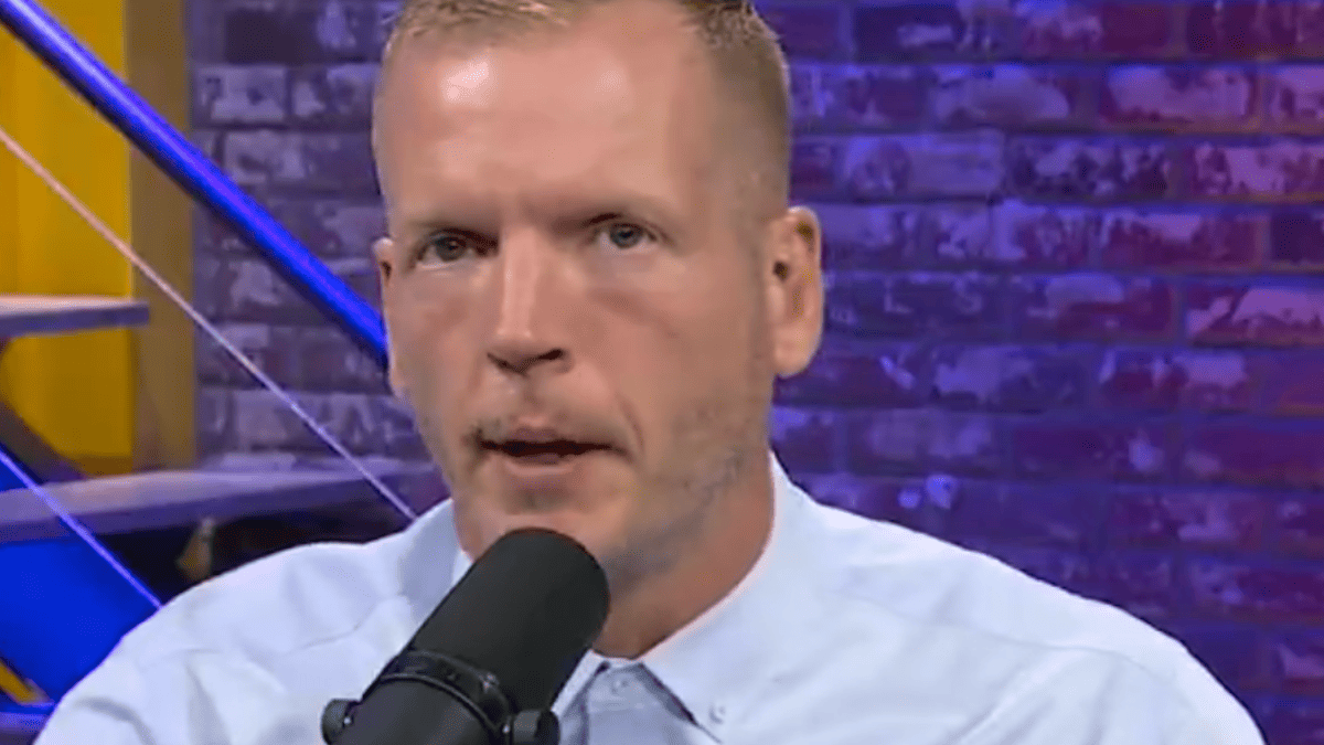 Chris Simms speaks into a microphone, wearing a light blue collared shirt, with a purple brick wall behind him.