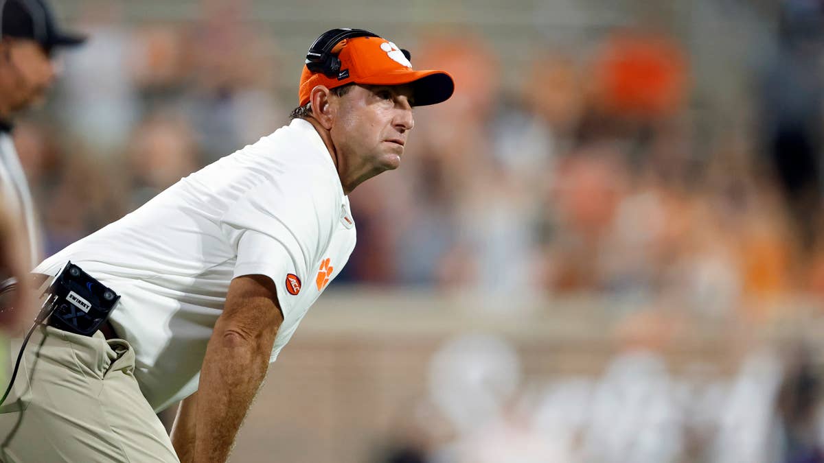Dabo Swinney in white Clemson shirt and orange hat with headphones, looking down.