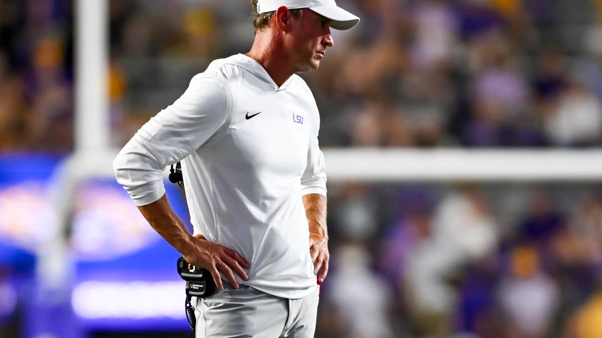 LSU coach in white hoodie and grey pants with hands on hips, looking to the side.