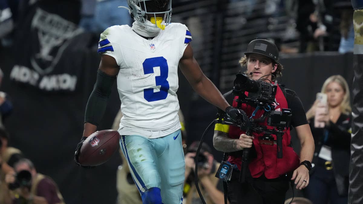 Cowboys player with number 3 holding a football, with a cameraman and fans in the background.