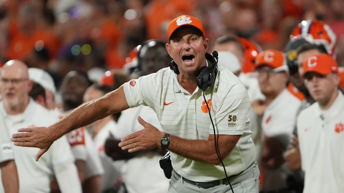 Dabo Swinney in a white polo and orange cap with Clemson logo, gesturing on the sideline.