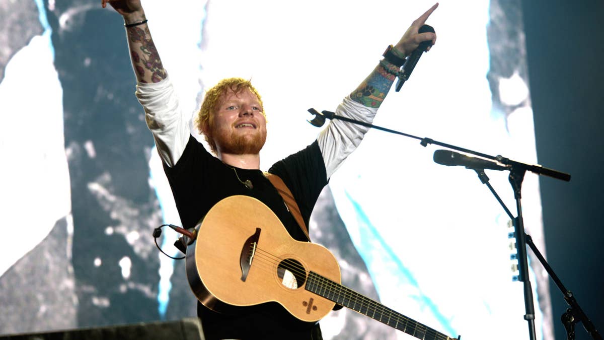 Ed Sheeran with arms raised, holding a guitar, wearing a black shirt and white sleeves.