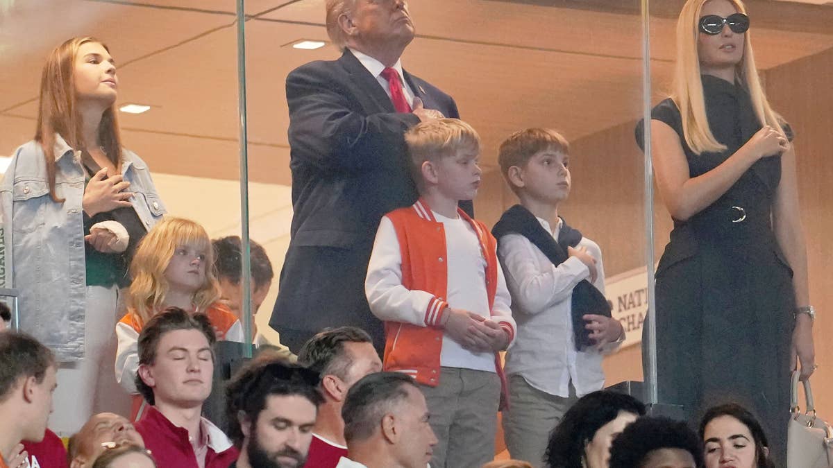 Donald Trump, Ivanka Trump, and children watch a football game, with Trump and Ivanka placing hands over their hearts.