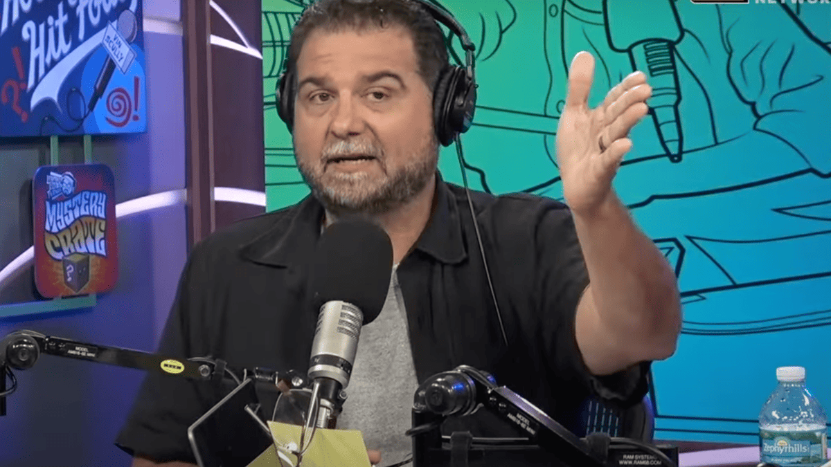 Dan Le Batard wearing a black shirt and headphones speaks into a microphone with a "Mystery Crate" sign behind him.