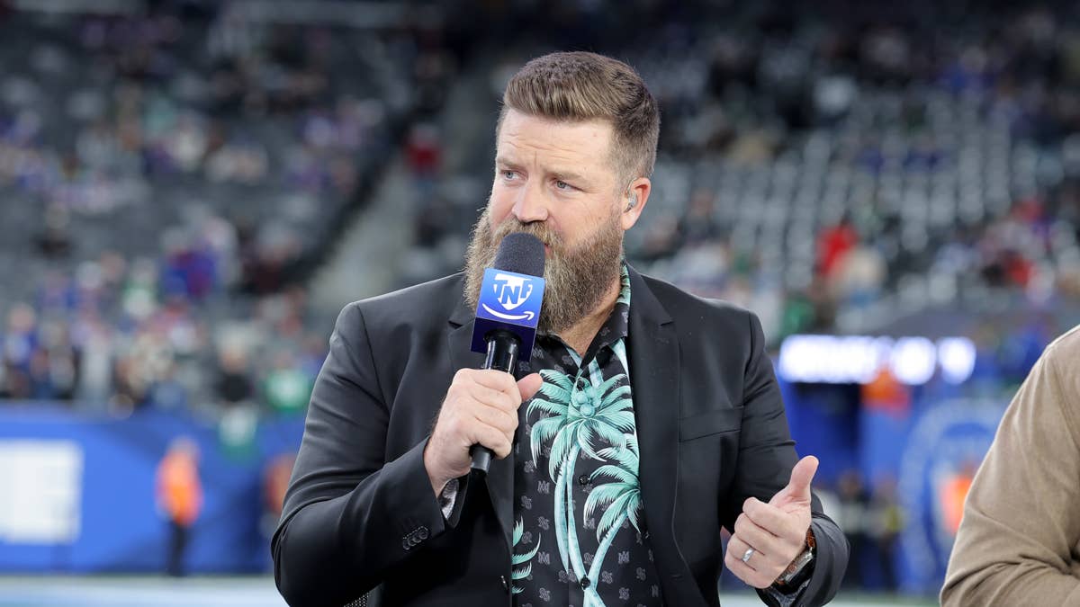 Ryan Fitzpatrick in a black suit and palm tree shirt holds a TNT microphone on a football field.