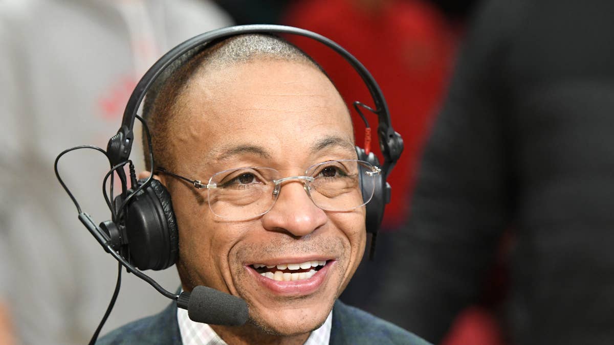 Gus Johnson wearing headphones and glasses smiles, with a microphone in front of him.