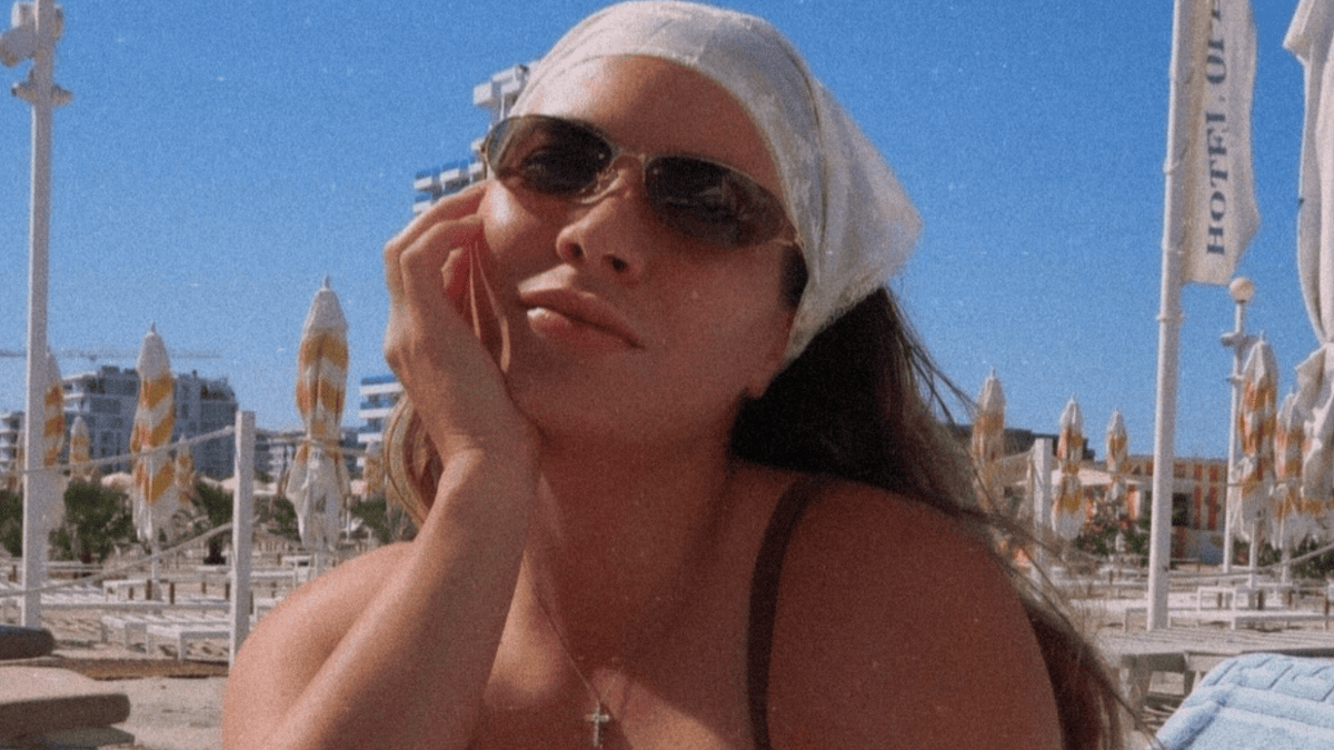 Gabriela Ruse in a swimsuit and sunglasses at the beach with umbrellas and hotel buildings in the background.