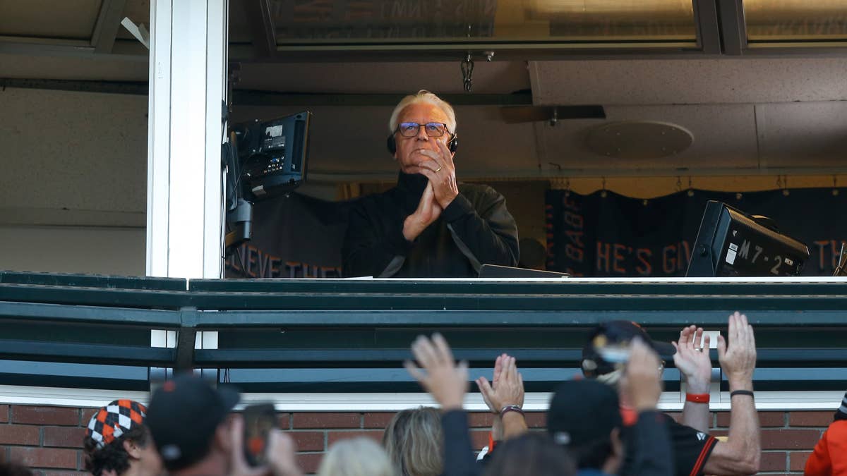 Duane Kuiper in broadcast booth with hands clasped, wearing glasses and a black jacket, with fans in foreground.