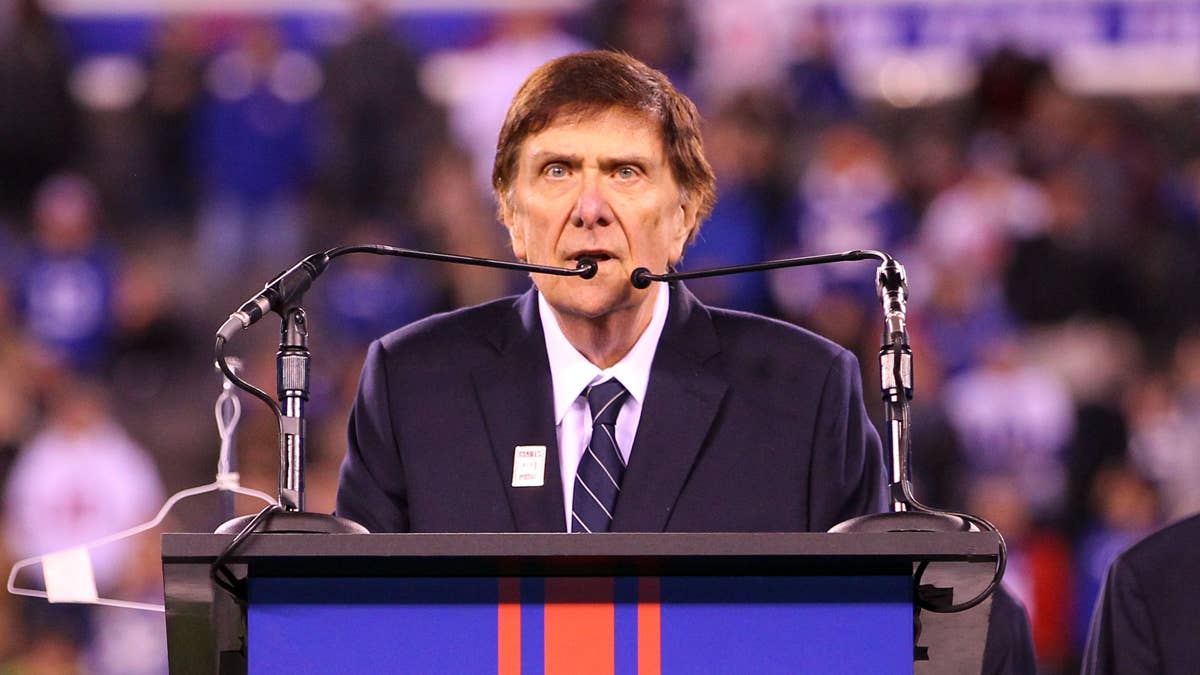 Ernie Accorsi at a podium in a navy suit and tie, speaking into microphones with a blurred crowd in the background.