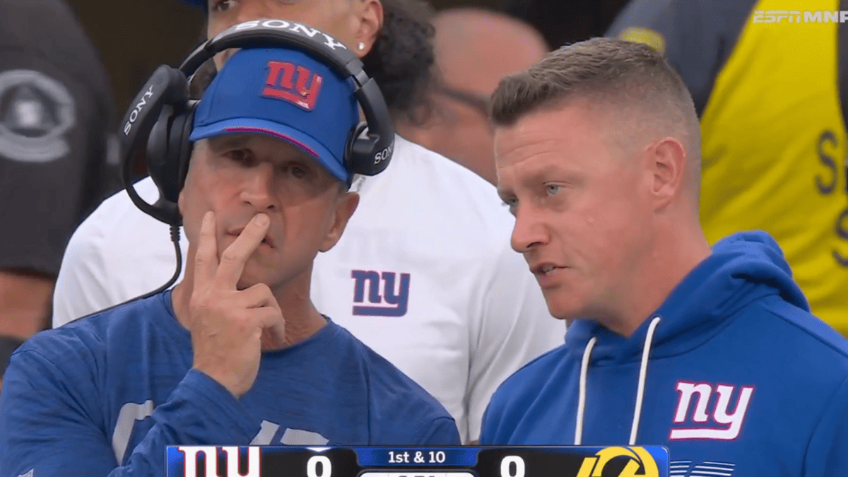 Giants coach with headphones and a blue NY Giants cap talks with another coach in a blue NY Giants hoodie.