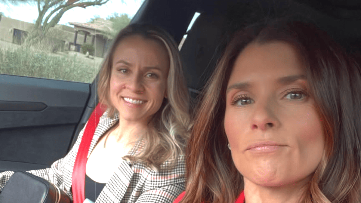 Danica Patrick and a friend smile in a car, wearing a plaid blazer and red seatbelt.