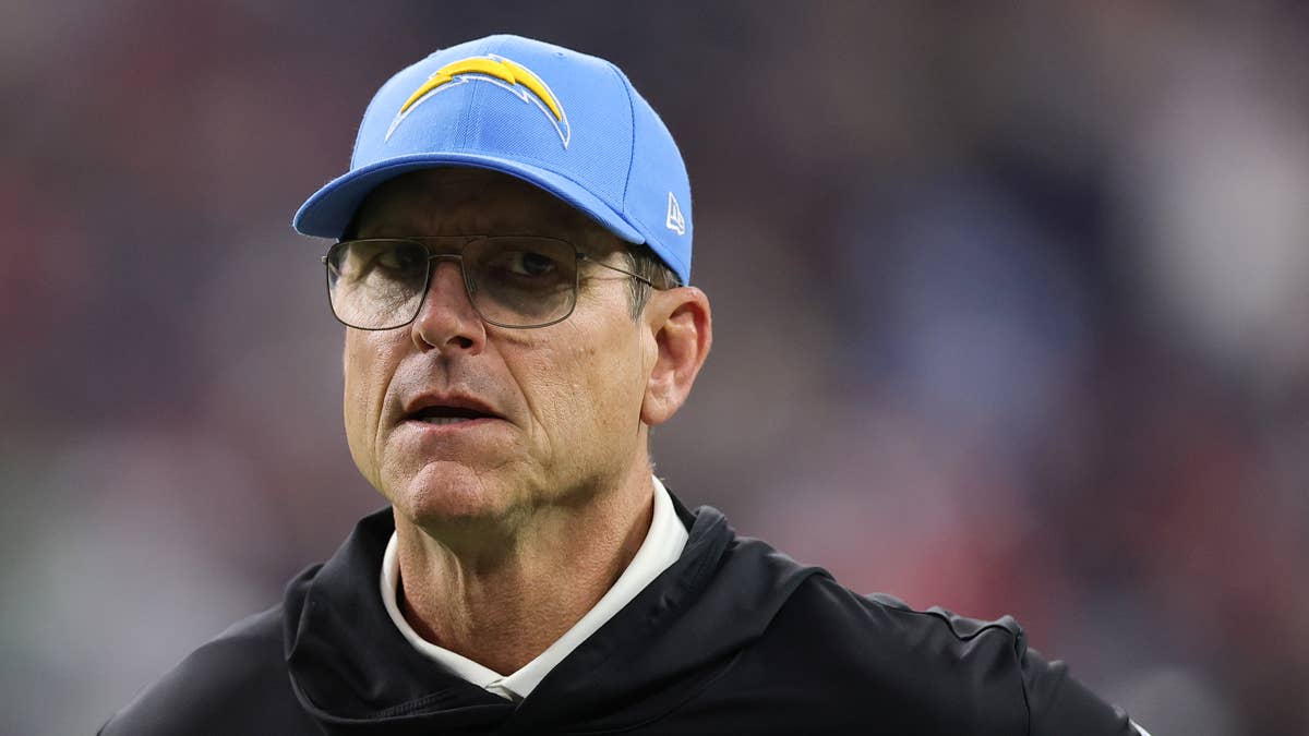 Jim Harbaugh wearing a Chargers cap and black jacket, looking concerned.