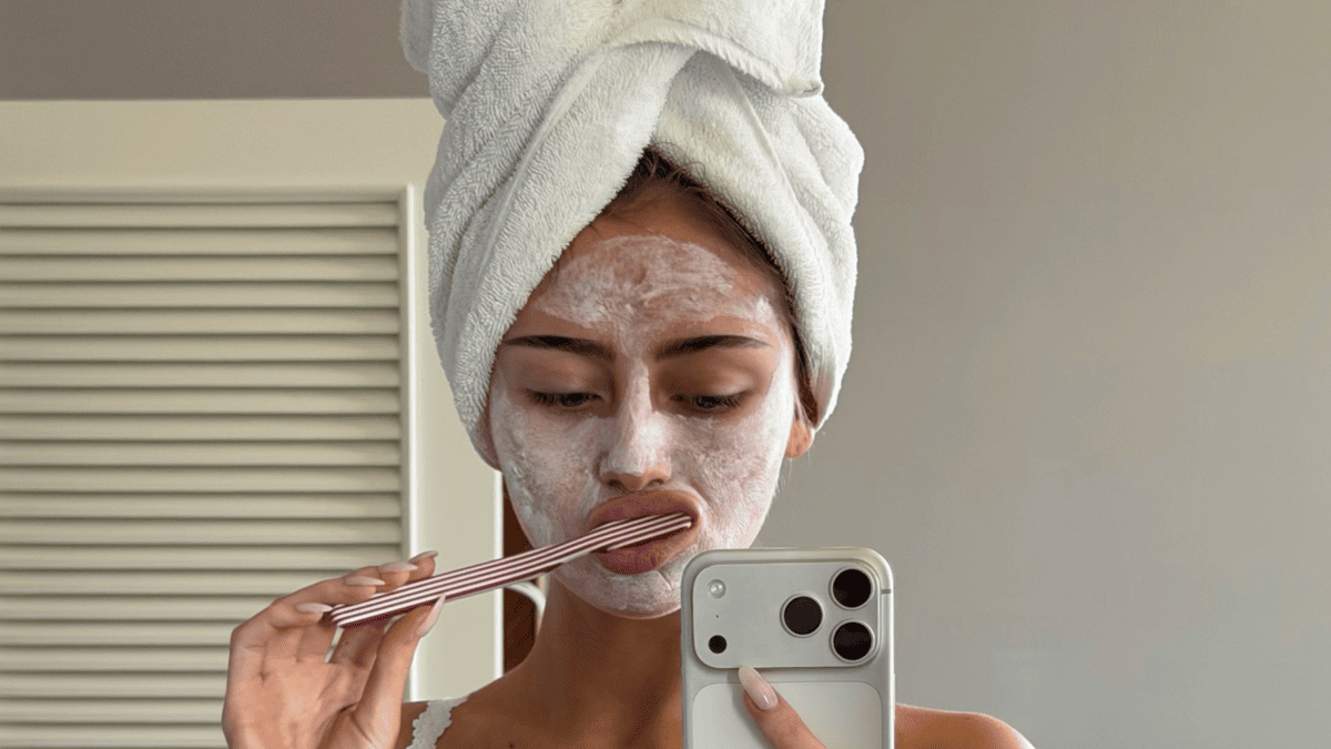 Leni Klum with a white towel on her head and a face mask on, holding a toothbrush and a phone.