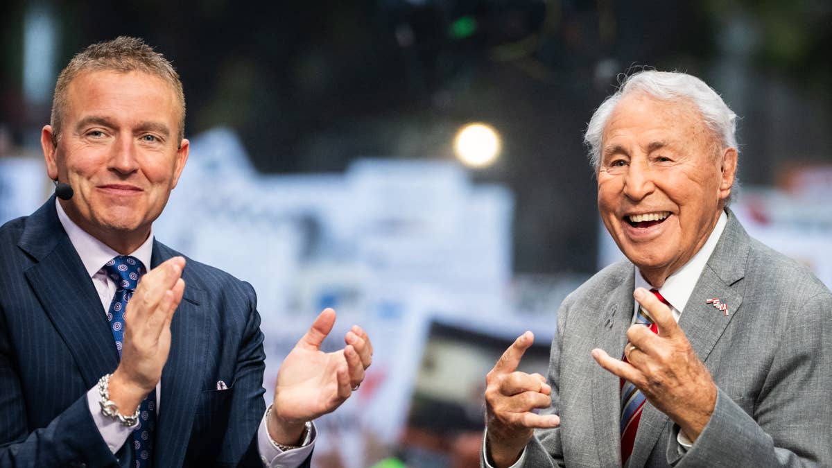Kirk Herbstreit in a pinstripe suit and blue tie, and Lee Corso in a grey suit and red tie, both smiling.