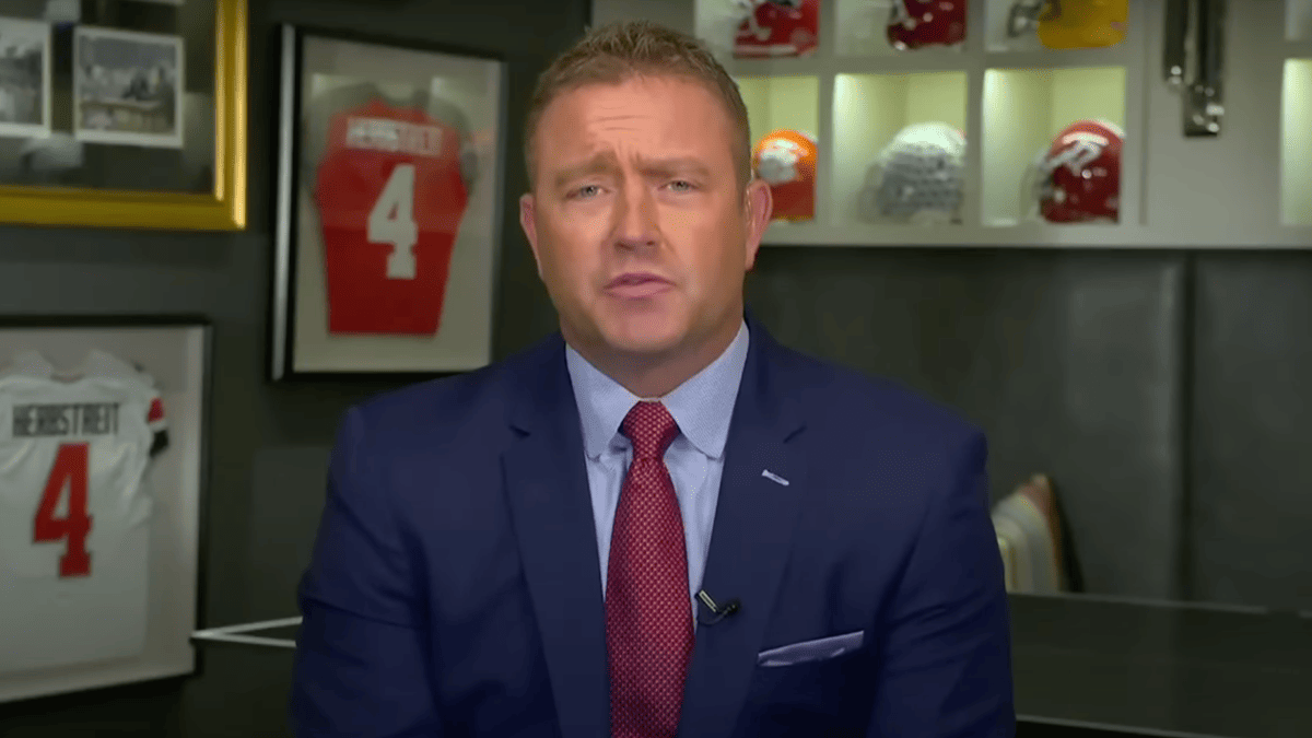 Kirk Herbstreit in a blue suit and red tie, with framed jerseys and football helmets behind him.