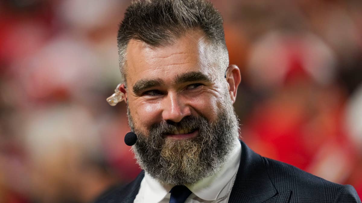 Jason Kelce in a suit with a microphone and earpiece, smiling with a beard.