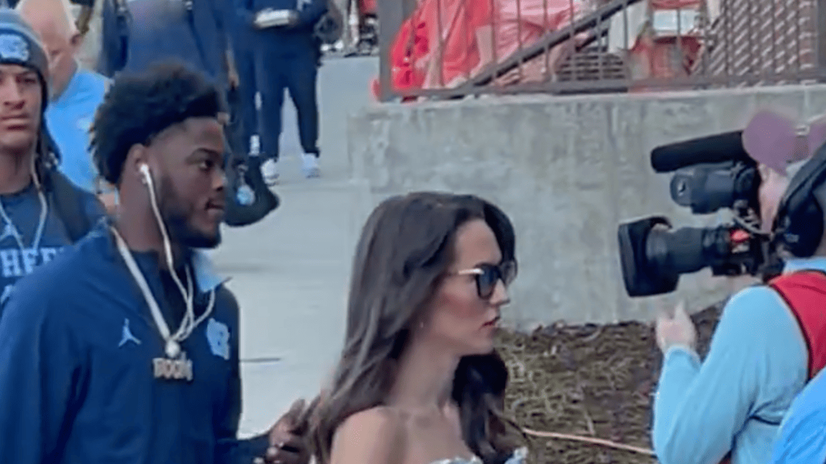 Jordon Hudson in a sparkly dress and a UNC player in a blue jacket with a "BOOM" necklace walk past a camera crew.