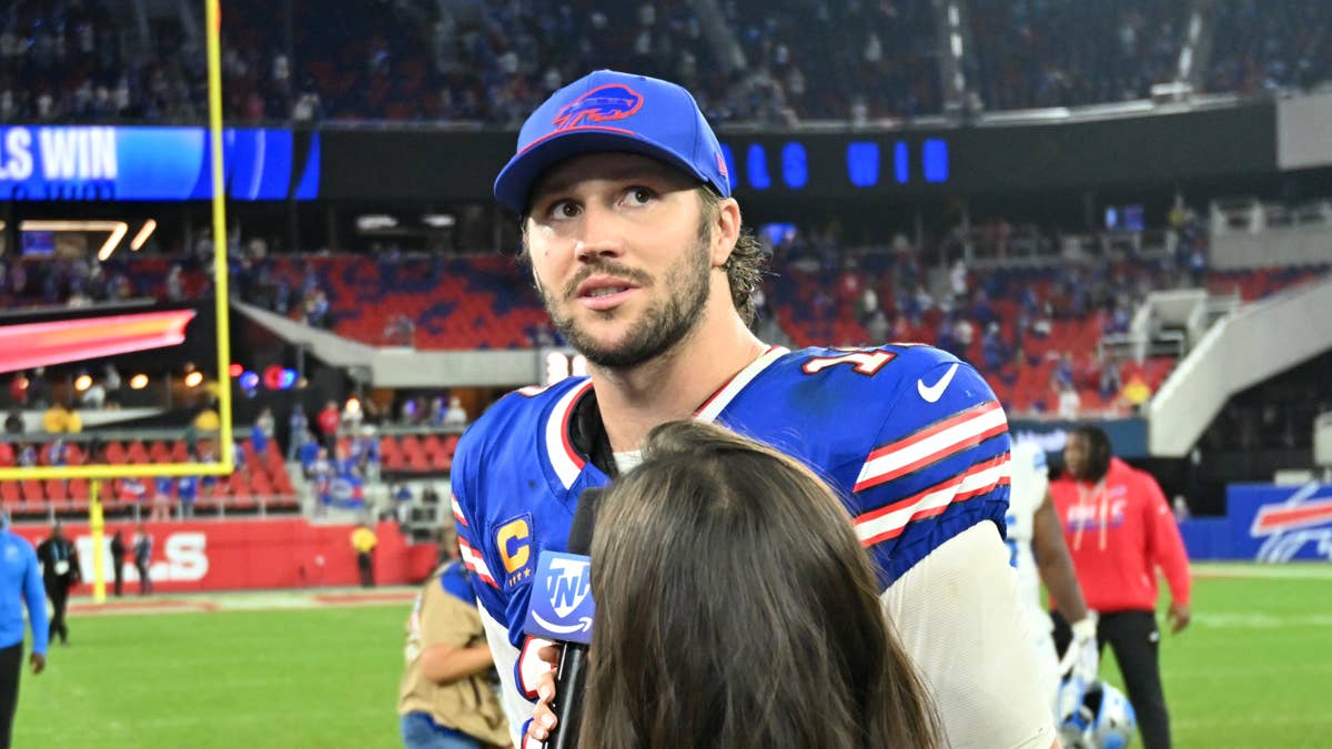 Josh Allen in a blue Bills jersey and cap speaks to a reporter on a football field.