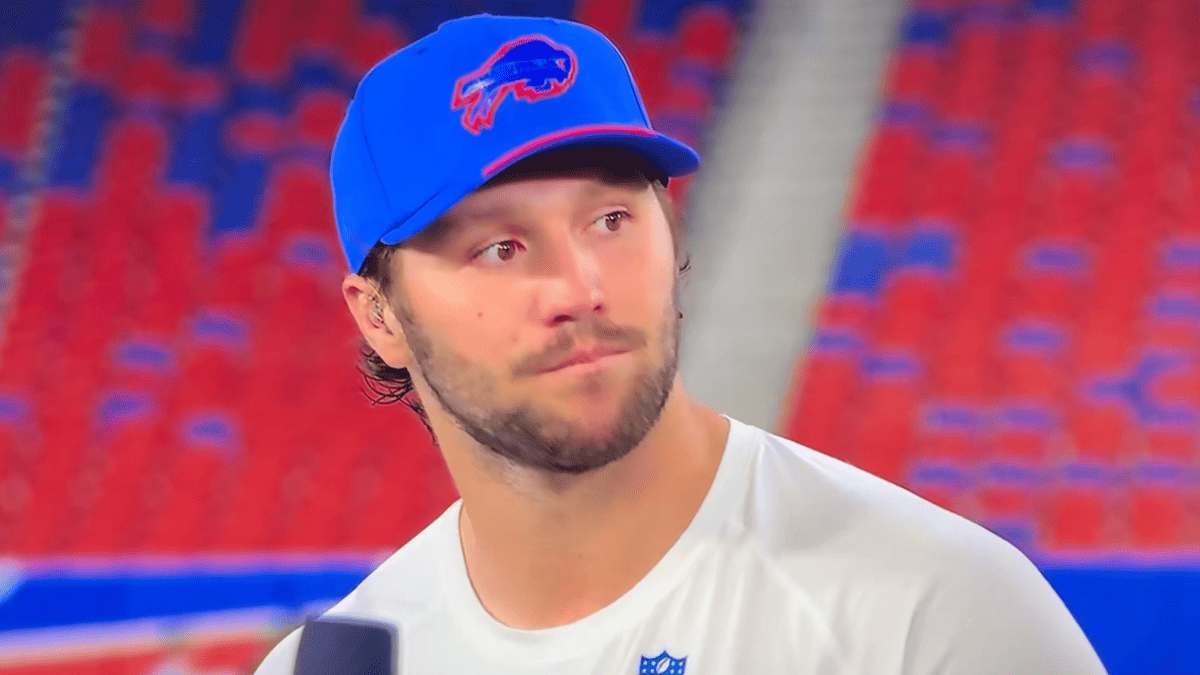 Josh Allen in a blue Bills hat and white shirt with NFL logo, looking off to the side.