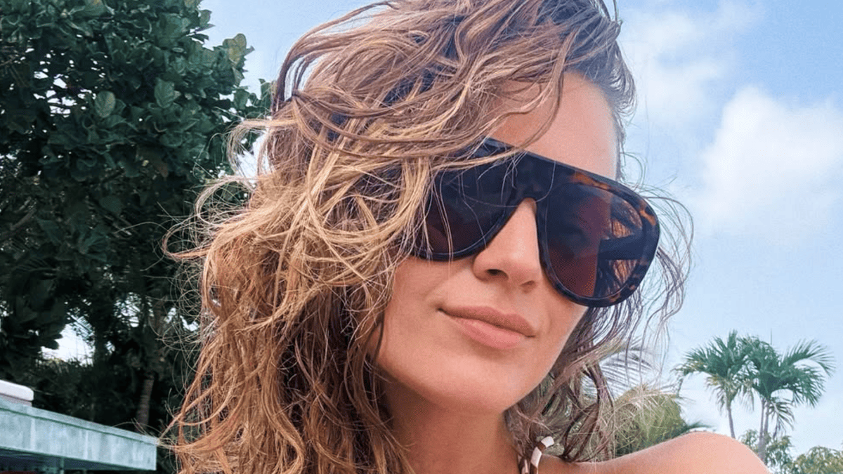Kelsea Ballerini in sunglasses and a swimsuit with wet hair and palm trees in the background.