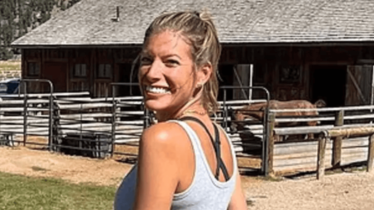 Jennifer Milburn in a grey tank top smiles in front of a wooden barn and corral with a horse.
