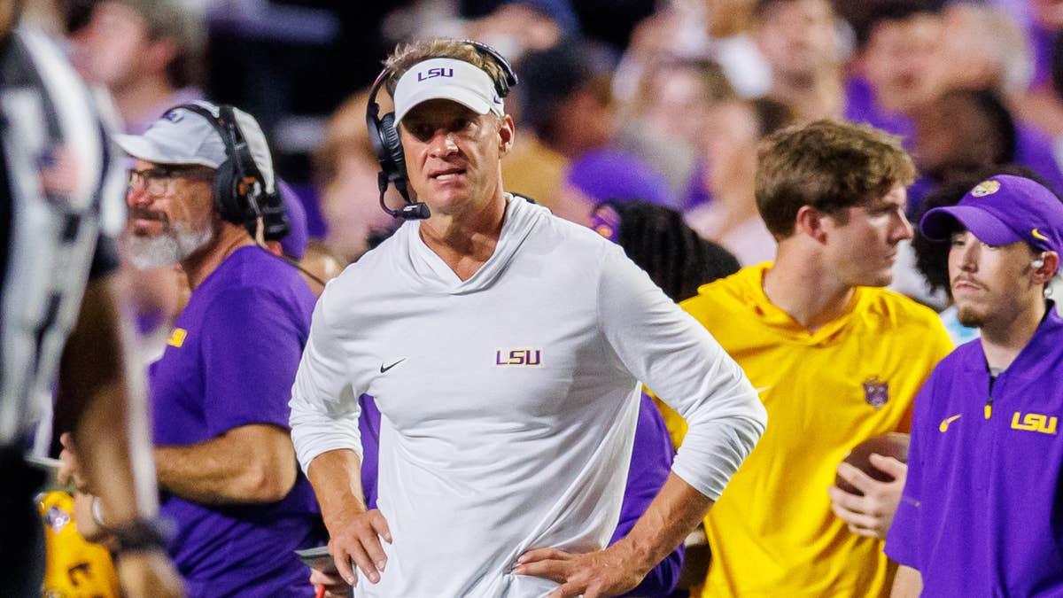 Lane Kiffin in a white LSU hoodie and visor on the sideline with other coaches.