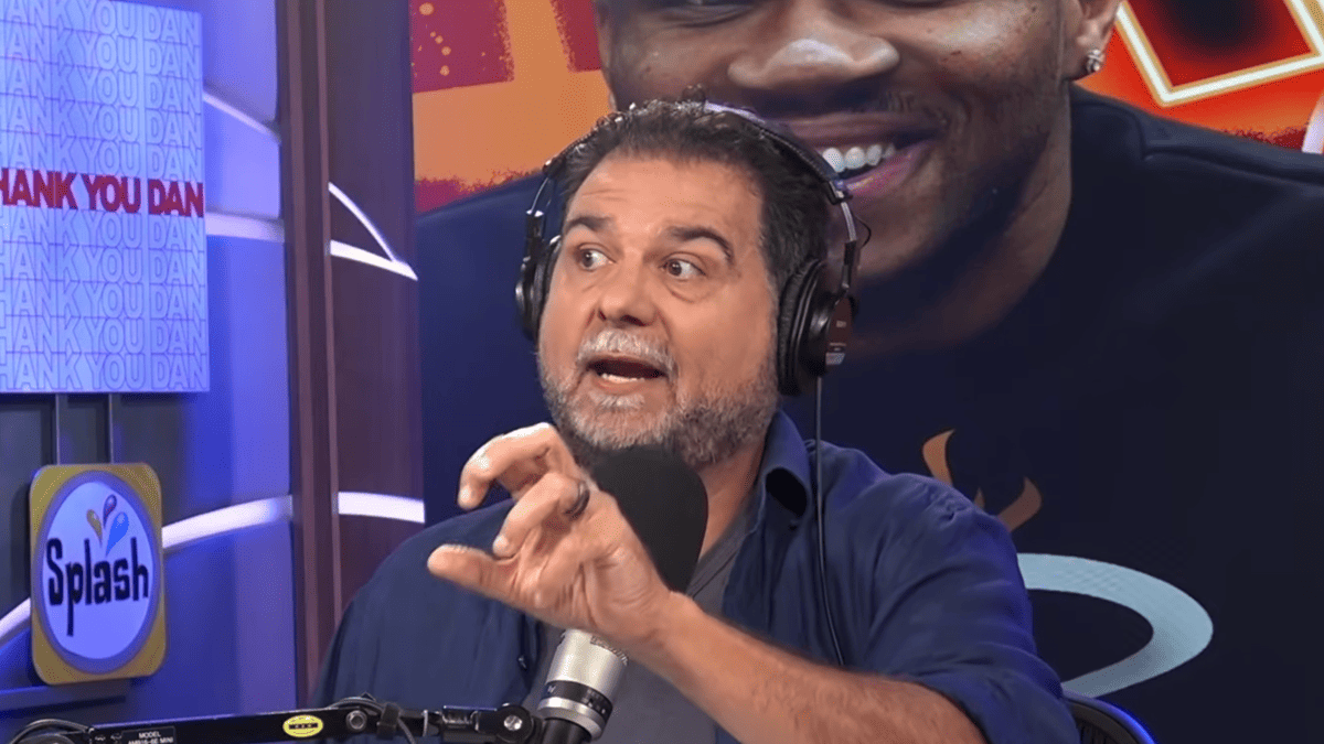 Dan Le Batard wearing headphones and a blue shirt gestures while speaking at a microphone, with a "Thank You Dan" sign behind him.