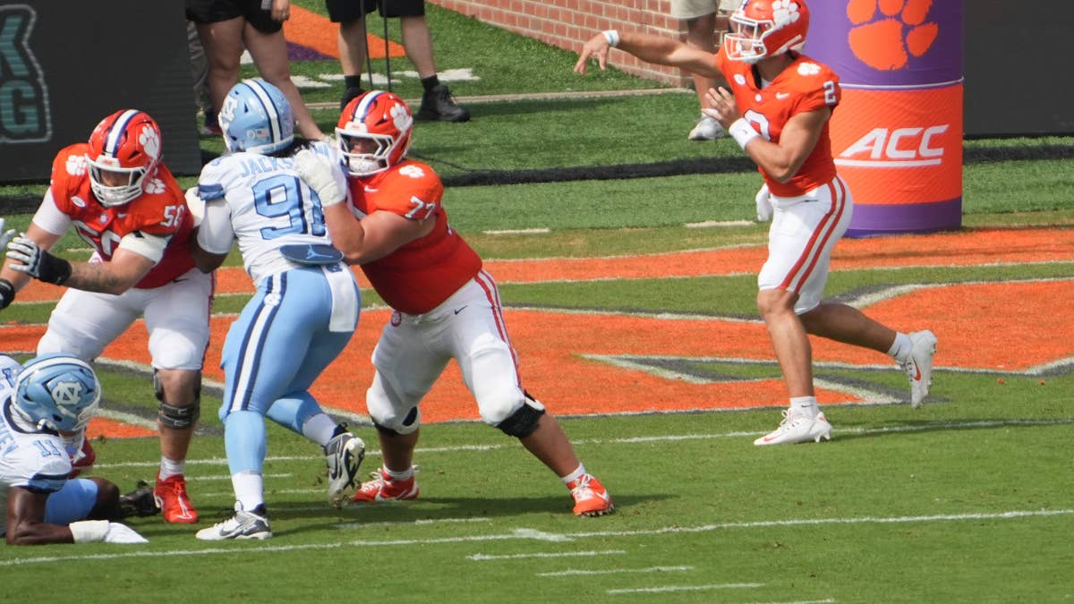 Clemson quarterback #2 throws a pass during a game against UNC, with players in orange and light blue uniforms on the field.