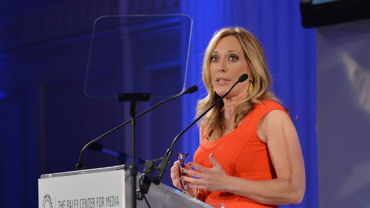 Linda Cohn speaks at a podium wearing an orange dress and large gold earrings.