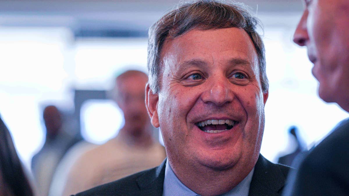 Michael Lombardi in a suit and tie smiles broadly while talking to another man.