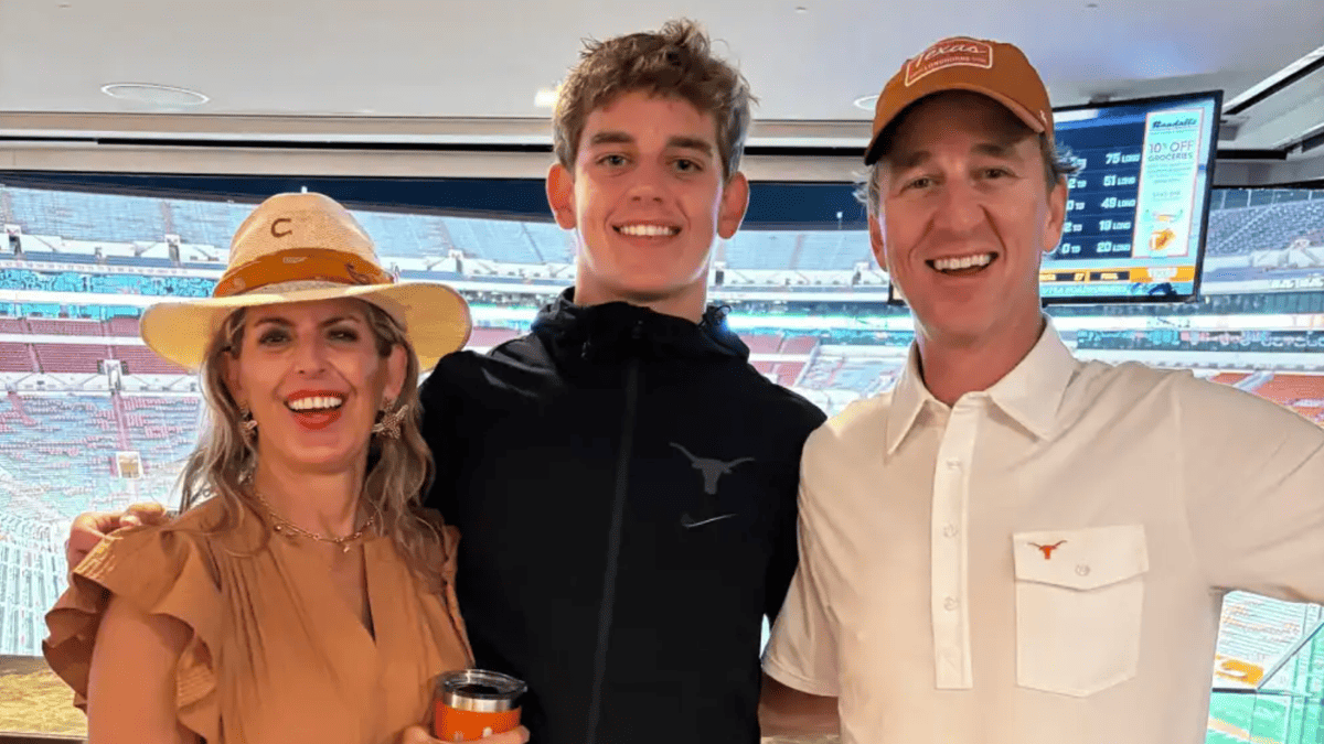 Arch Manning smiles with his family at a football game, wearing a black jacket with a Texas Longhorns logo.