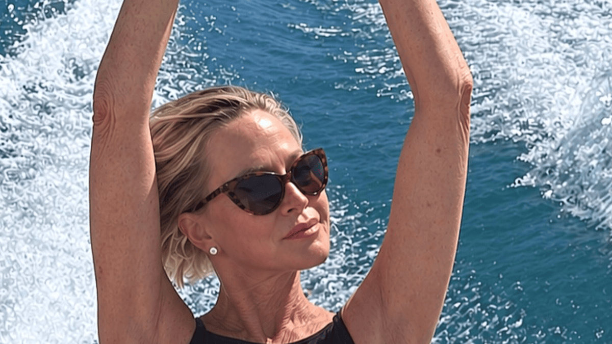 Melanie Griffith in sunglasses and a black swimsuit with arms raised on a boat with blue water.