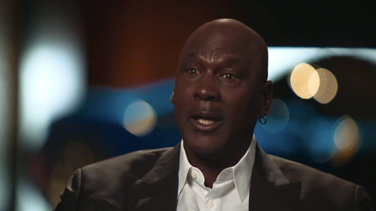 Michael Jordan in a suit and white shirt with a hoop earring, speaking with blurred lights in the background.