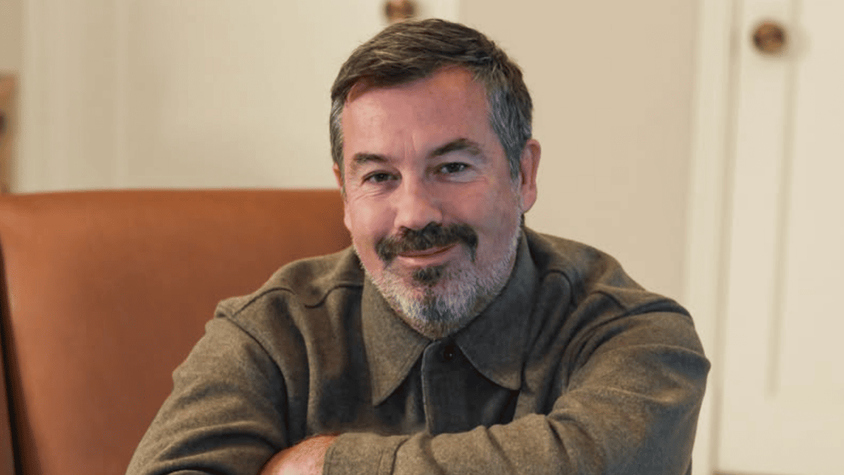 Duncan Sheik smiles, wearing a brown collared shirt, seated in a brown chair.