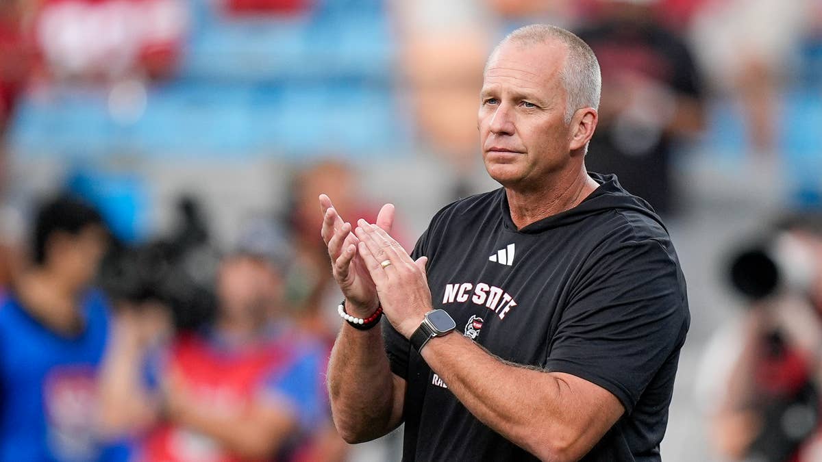NC State coach Dave Doeren in black shirt with NC State logo claps hands on sideline.