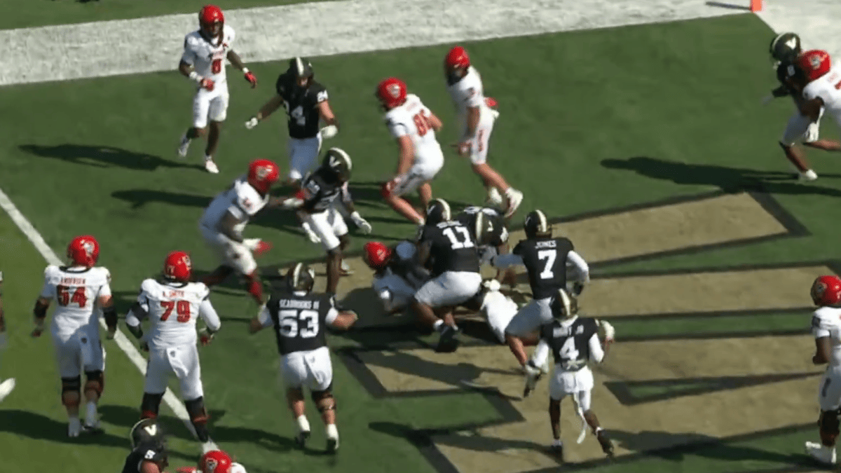 NC State players in white uniforms with red helmets and Vanderbilt players in black uniforms with gold helmets scramble for a fumble on the field.
