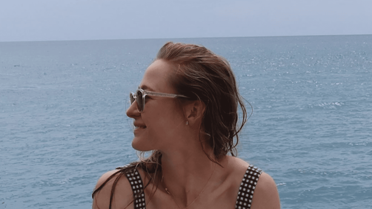 Nelly Korda in a black and white checkered swimsuit and sunglasses smiles by the ocean in Greece.