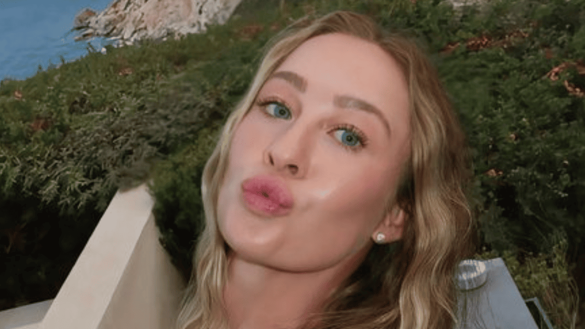 Nelly Korda pouts in a swimsuit with blue eyes and blonde hair, with green bushes and blue water in the background.
