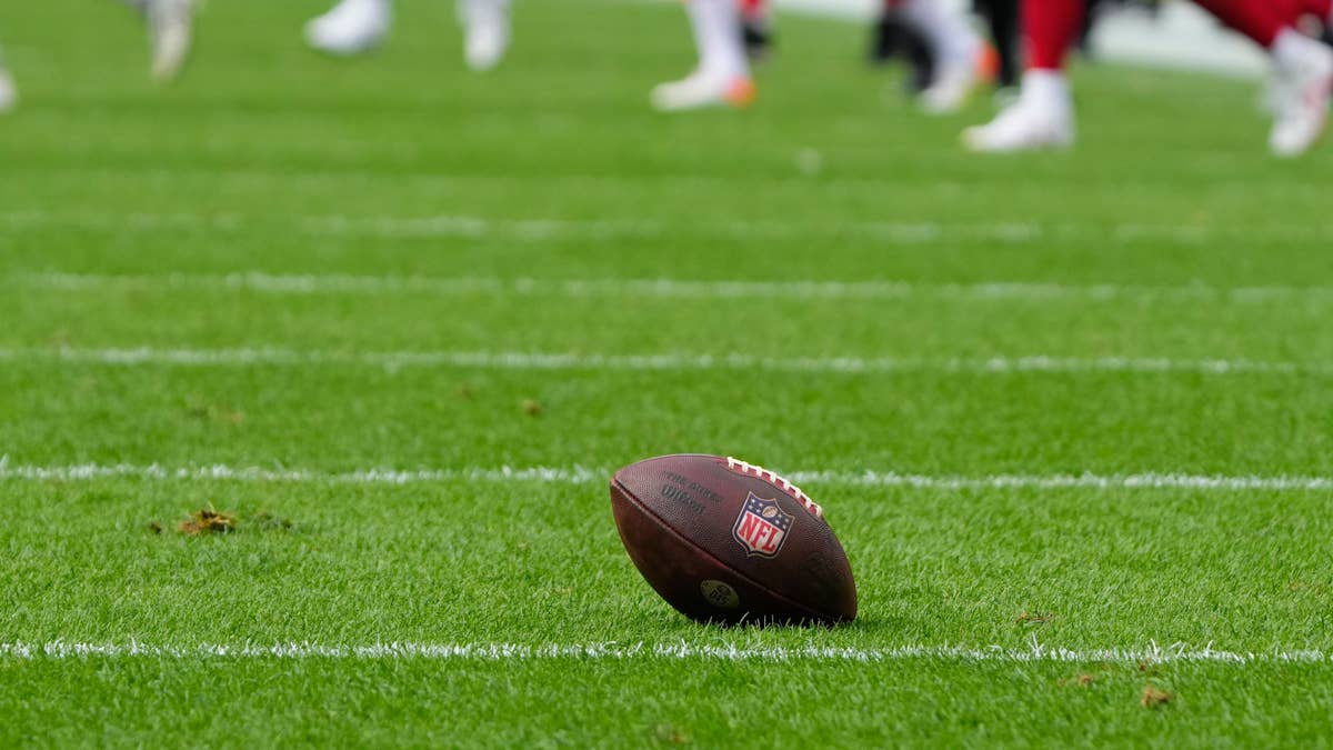 An NFL football rests on a green field with white yard lines. Blurred players in white and red uniforms are visible in the background.