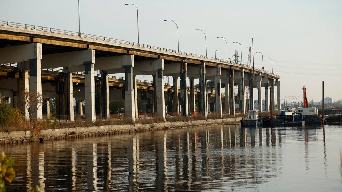 A concrete overpass with multiple lanes of traffic runs over a calm river with barges and construction equipment.