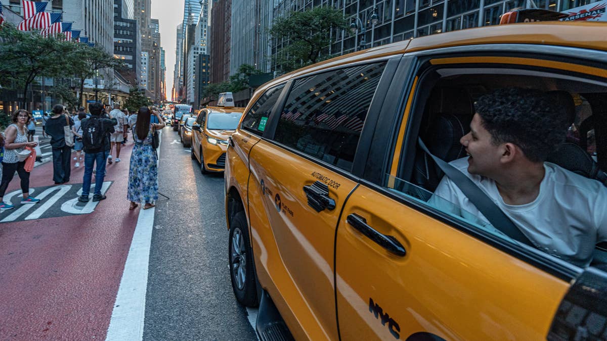 A young male NYC taxi driver in a white t-shirt smiles as he looks back from the driver's seat of a yellow taxi.